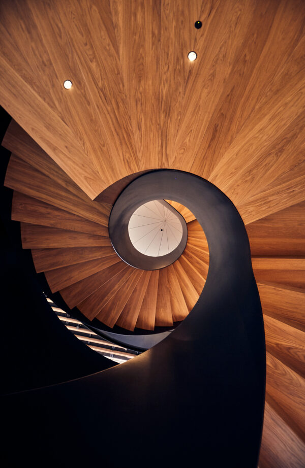 Upward view of a sculptural wooden spiral staircase with a smooth black handrail, creating a dramatic swirling pattern toward a circular skylight.